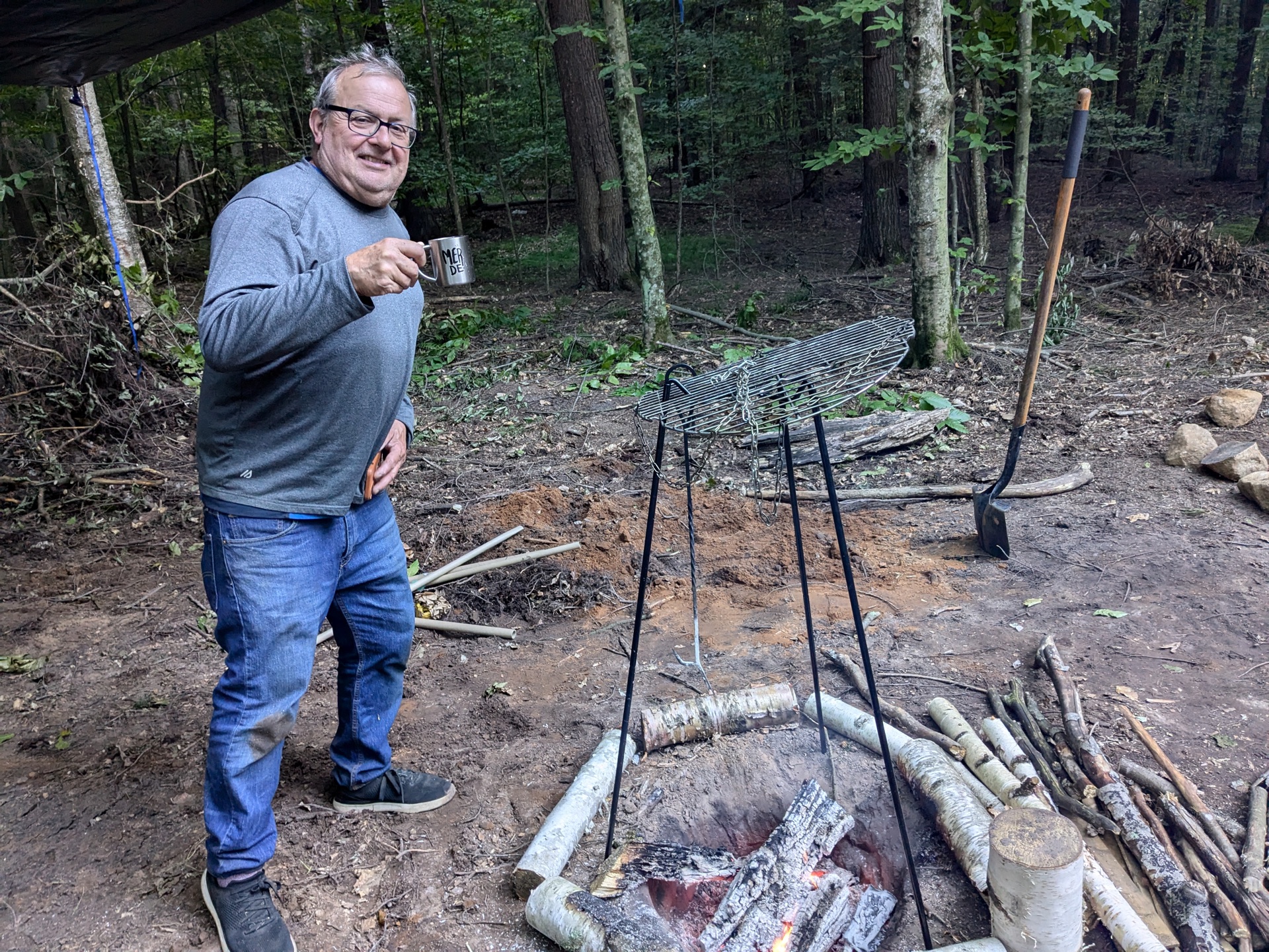 A camper raising a metal mug beside the fire pit and swing-arm grill